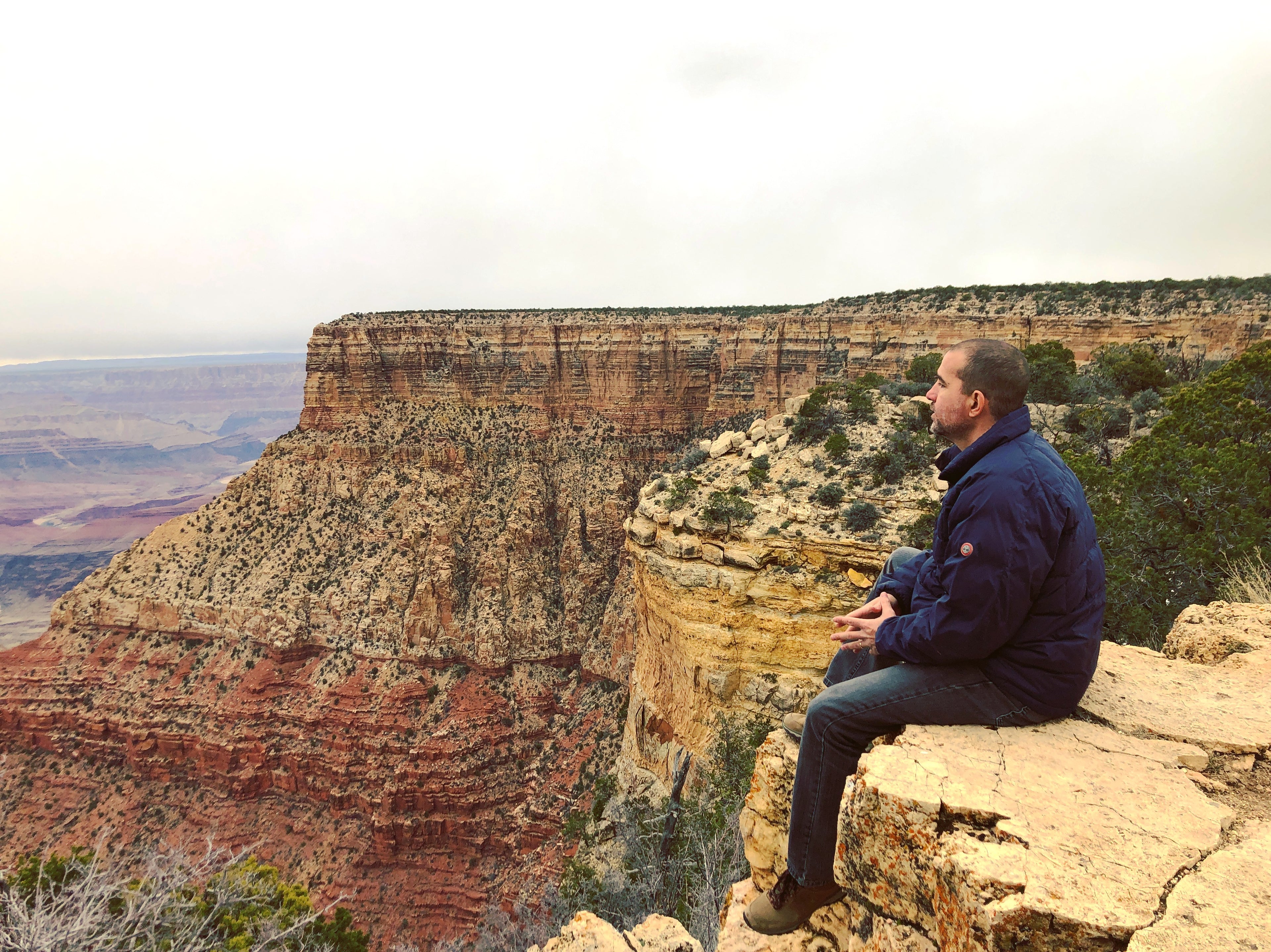 Phil (ANAAMALY) sitting on a rocky edge overlooking the vast landscape  of the Grand Canyon with cliffs and greenery.